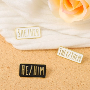 Detailed close-up of SHE/HER, THEY/THEM, and HE/HIM enamel pronoun pins on a wooden surface