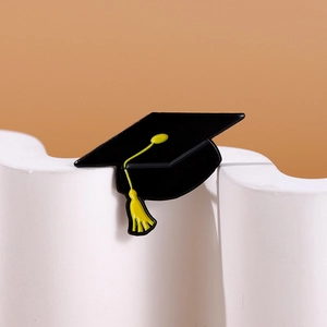 Detail view of a black enamel graduation cap brooch with a yellow tassel on a textured base