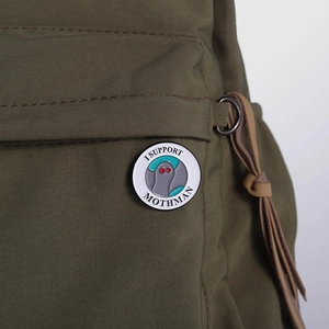 Detail view of round Mothman enamel pin with text attached to a green nylon backpack