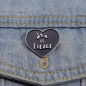 Detail of a black heart enamel brooch with She IS FIERCE text on blue denim fabric
