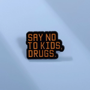 Macro shot of a black enamel pin with orange text saying SAY NO TO KIDS, DRUGS