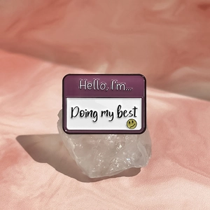 Gros plan du pin's émaillé Hello I'm Doing My Best sur un fond rose pastel doux
