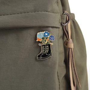 Enamel pin featuring blue flowers in a black leather boot pinned to an olive backpack