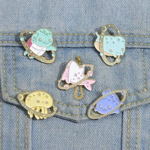 Set of five cartoon space animal enamel pins displayed on blue denim fabric background