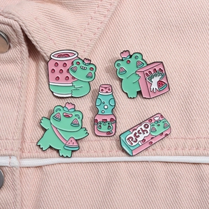 Cinq broches en émail grenouille et pastèque présentées sur un tissu en jean rose