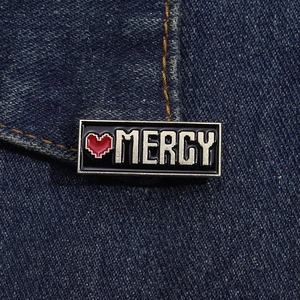 Dettaglio macro di una spilla smaltata con cuore pixelato Mercy su tessuto denim blu