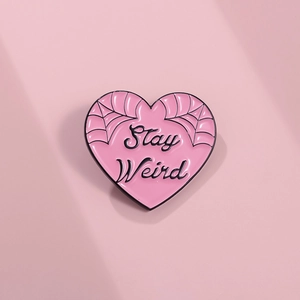 Close-up of pink heart enamel pin with Stay Weird slogan and spider web detail