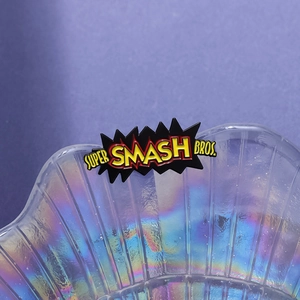 Macro shot of Super Smash Bros enamel pin with gradient text on a glossy shell tray