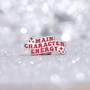 Rode emaille pin met Main Character Energy tekst op een zilveren glitterachtergrond
