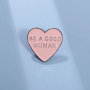 Spilla smaltata cuore rosa con testo nero BE A GOOD HUMAN su sfondo azzurro chiaro