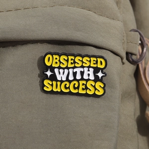Close-up of Obsessed with Success enamel pin badge on green canvas bag fabric