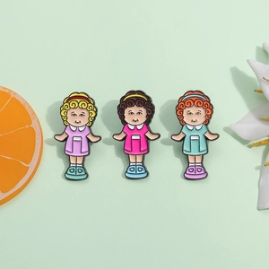 Three-piece cartoon girl enamel pins in purple, pink, and blue dresses on a green backdrop