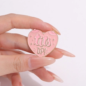 Close-up of pink heart enamel brooch featuring tits UP! text and gold metal outline