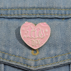 Pink heart enamel pin with tits UP! text on blue denim fabric background