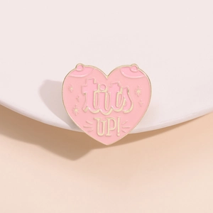 Pink heart-shaped enamel pin with gold metal trim and 'tits UP!' lettering decoration