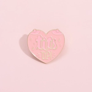 Detail of pink heart enamel pin featuring Tits Up text and golden metallic edge design