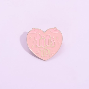 Detailed close-up of heart-shaped pink enamel pin with tits up text and gold metal edges