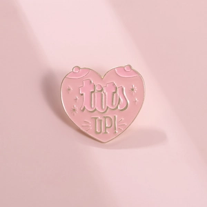Detail shot of heart enamel pin with 'tits UP!' lettering and golden outlines