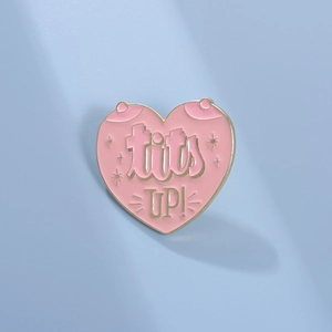 Detailed view of pink heart enamel pin with gold Tits Up text on blue background