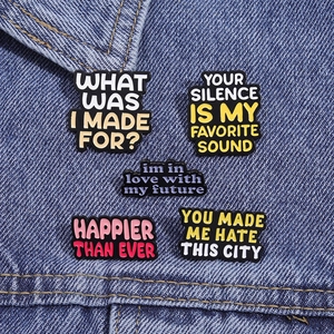 Set of 5 enamel pins with colorful quotes like Happier Than Ever on denim background