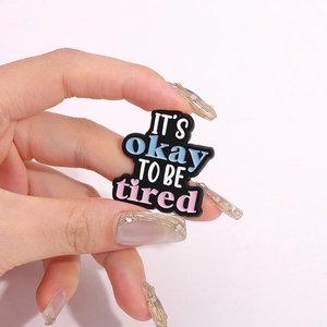 Handgehaltener Pin mit dem Text IT'S OKAY TO BE TIRED und einem kleinen Herz-Detail