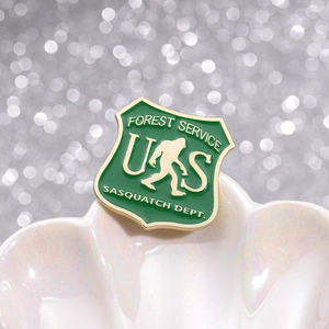 Funny green Forest Service Sasquatch Dept enamel pin with gold lettering on white shell base