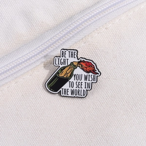 Detail of creative enamel frog pin featuring Be the light text on a white canvas bag