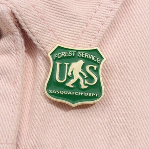 Green metal shield enamel pin featuring US Forest Service Sasquatch Dept on pink fabric