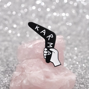 Minimalist black Karma boomerang enamel pin displayed on pink natural quartz stones
