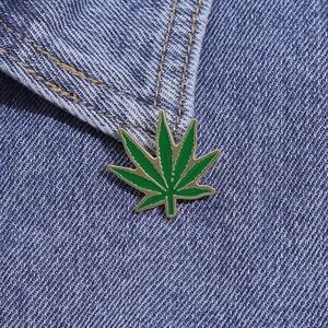 Macro view of green enamel maple leaf pin on blue denim fabric with golden edges