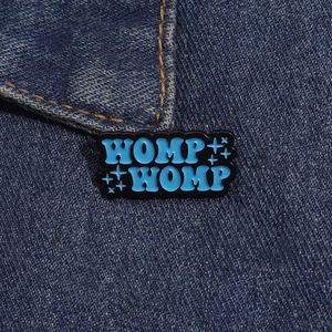 Primo piano di una spilla smaltata blu WOMP WOMP sul tessuto di jeans scuro