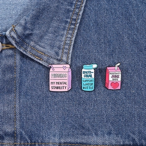 Three colorful enamel pins with humorous slogans pinned on blue denim jacket fabric