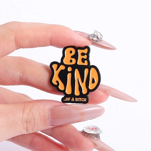 Detail of creative orange enamel pin with 'BE KIND ..OF A BITCH' text on black metal
