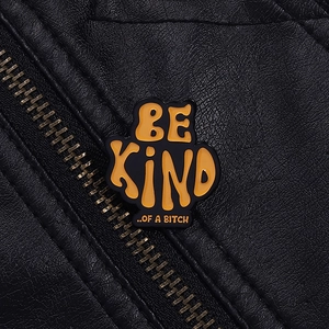 Close-up of a black enamel pin with orange BE KIND OF A BITCH text on a leather jacket