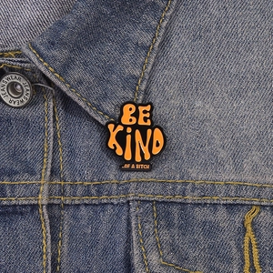 Vintage orange and black enamel pin with Be Kind Of A Bitch text on denim jacket