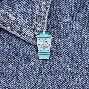 Detailed view of an enamel pin shaped like a drink cup pinned to blue denim fabric