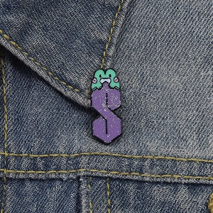 Dettaglio di una spilla smaltata a rana verde con S glitterata viola su denim