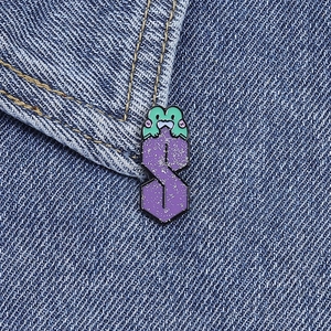 Dettaglio di una spilla a forma di rana con lettera S glitter viola su giacca in denim