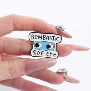 Dettaglio della spilla in smalto blu con scritta Bombastic Side Eye tenuta in mano su bianco