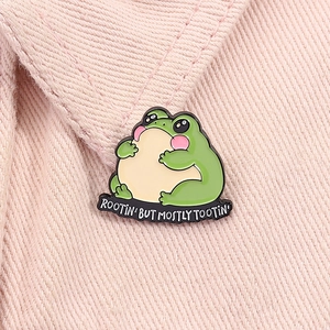 Detail of cute green frog enamel pin with Rootin' But Mostly Tootin' text on pink denim
