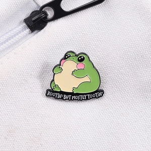 Close-up of green frog enamel pin with Rootin' But Mostly Tootin' slogan on white canvas