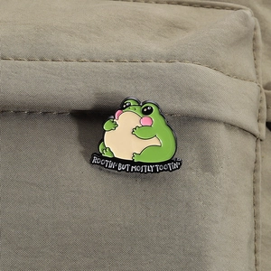 Close-up of a cute green frog enamel pin on a backpack with the slogan Rootin' Tootin'