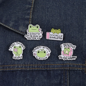 Detail showing five cute green frog enamel pins with text on blue denim fabric
