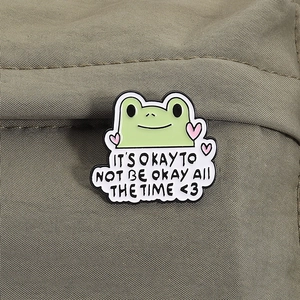 Detail of cute green frog enamel pin on khaki canvas bag with motivational quote text