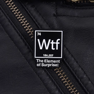 Détail d'une broche en émail noir avec l'élément Wtf et texte The Element of Surprise humour