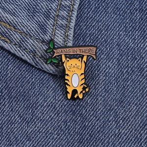 Détail de la broche chat en émail orange avec texte Hang In There sur tissu denim