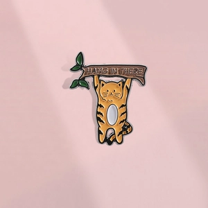 Détail d'un pin's en émail avec un chat orange et le texte Hang In There sur une branche