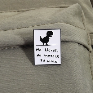 Detail of white enamel rectangular brooch with black dinosaur and Spanish text on fabric