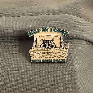 Detail of enamel raccoon brooch with Hop In Loser text on green canvas background