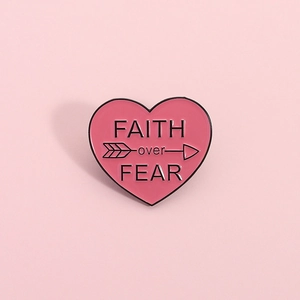 Broche en émail cœur rose avec texte Faith over Fear sur un fond rose pastel